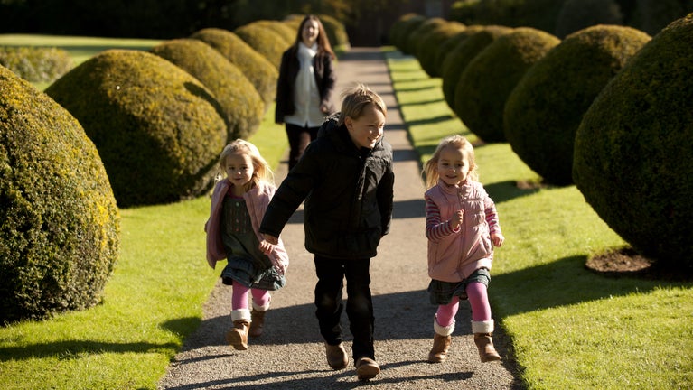 Three children walking along path lined with topiary while mother walks behind